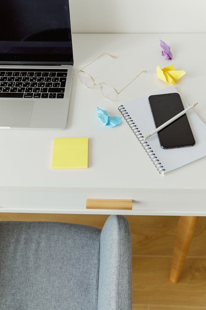 High-angle view of a minimalist desk with laptop, smartphone, notebook, and eyeglasses.