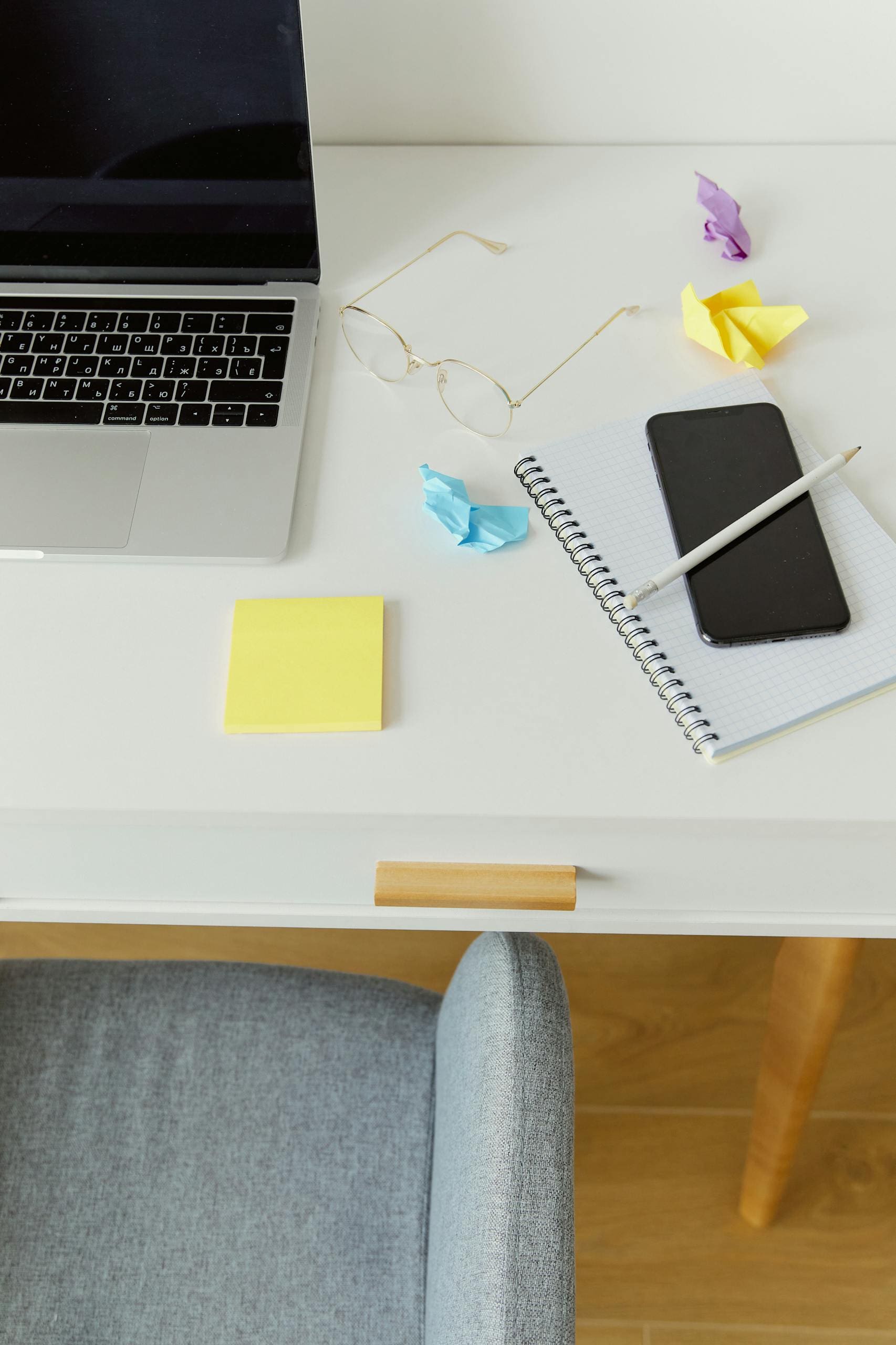 High-angle view of a minimalist desk with laptop, smartphone, notebook, and eyeglasses.