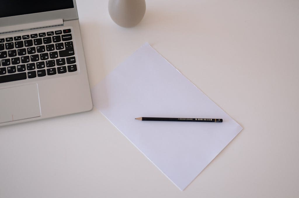 Top view of a minimalist workspace with laptop, paper, and pencil on a white table.